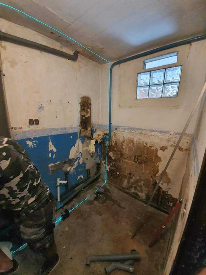 Łazienka w trakcie remontu z widocznymi uszkodzeniami ścian, instalacją hydrauliczną i poziomnicą laserową wyznaczającą linie, fragment osoby w ubraniu moro w lewym dolnym rogu.