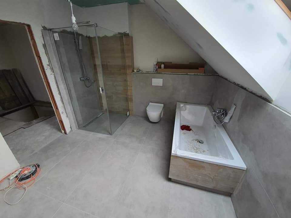 Łazienka w trakcie remontu na poddaszu: szara glazura podłogowa, kabina prysznicowa ze szklanymi drzwiami, biała wanna obudowana drewnopodobną płytą, wisząca toaleta, narzędzia glazurnicze...