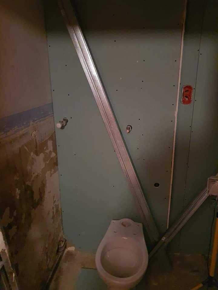 Wnętrze łazienki w trakcie remontu: częściowo zamontowana płyta gipsowo-kartonowa, widoczne profile metalowe, stara ściana z odpadającym tynkiem i stojący pisuar.