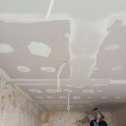 Mok-Bud remonty i wykończenia wnętrz - Pracownik szpachluje sufit z płyt kartonowo-gipsowych, widoczne kable wystające z sufitu i ściany z resztkami tapet.