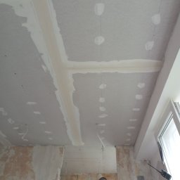 Mok-Bud remonty i wykończenia wnętrz - Sufit z płyt kartonowo-gipsowych w trakcie montażu, widoczne szpachlowane łączenia i otwory po wkrętach, ściany w stanie surowym, fragment okna i sylwetka pracownika.