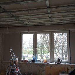 Mok-Bud remonty i wykończenia wnętrz - Wnętrze w trakcie remontu: widoczna drabina z farbami, okno z widokiem na drzewa, sufit z metalową konstrukcją pod płyty gipsowe, ściany częściowo wyszpachlowane, bałagan na parapecie i podłodze.