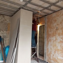 Mok-Bud remonty i wykończenia wnętrz - Montaż płyty karton gips na metalowym stelażu sufitowym w pomieszczeniu z częściowo otynkowanymi ścianami, widoczna drabina i zwisające kable elektryczne.