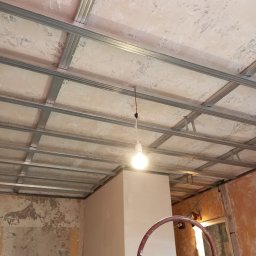 Mok-Bud remonty i wykończenia wnętrz - Niedokończony sufit podwieszany z metalowym stelażem i prowizorycznym oświetleniem żarówką, częściowo otynkowane ściany w tle, widoczny fragment drabiny malarskiej.