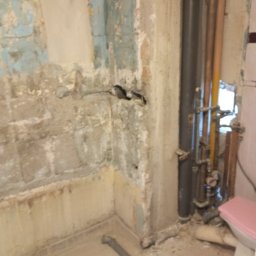 Mok-Bud remonty i wykończenia wnętrz - Zdjęcie przedstawia wnętrze łazienki w trakcie remontu, z widocznymi fragmentami starej, zniszczonej ściany z pozostałościami farby, odsłoniętymi rurami i instalacją wodną oraz fragmentem toalety...