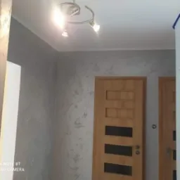 Ściana w pokoju wykończona dekoracyjną szarą farbą strukturalną, widoczne dwie drewniane drzwi z przeszkleniami, oświetlenie sufitowe z trzema reflektorami.