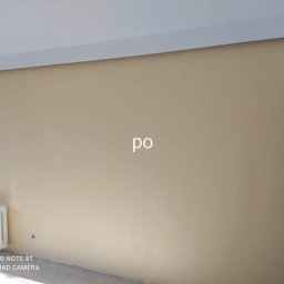 Mok-Bud remonty i wykończenia wnętrz - Ściana pomalowana na beżowo z białym grzejnikiem i listwą przysufitową, widoczny napis 'po' na ścianie.