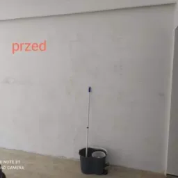 Pokój przed remontem: surowa ściana, widoczna niedoskonałość powierzchni, mop w wiadrze i napis 'przed' na ścianie.