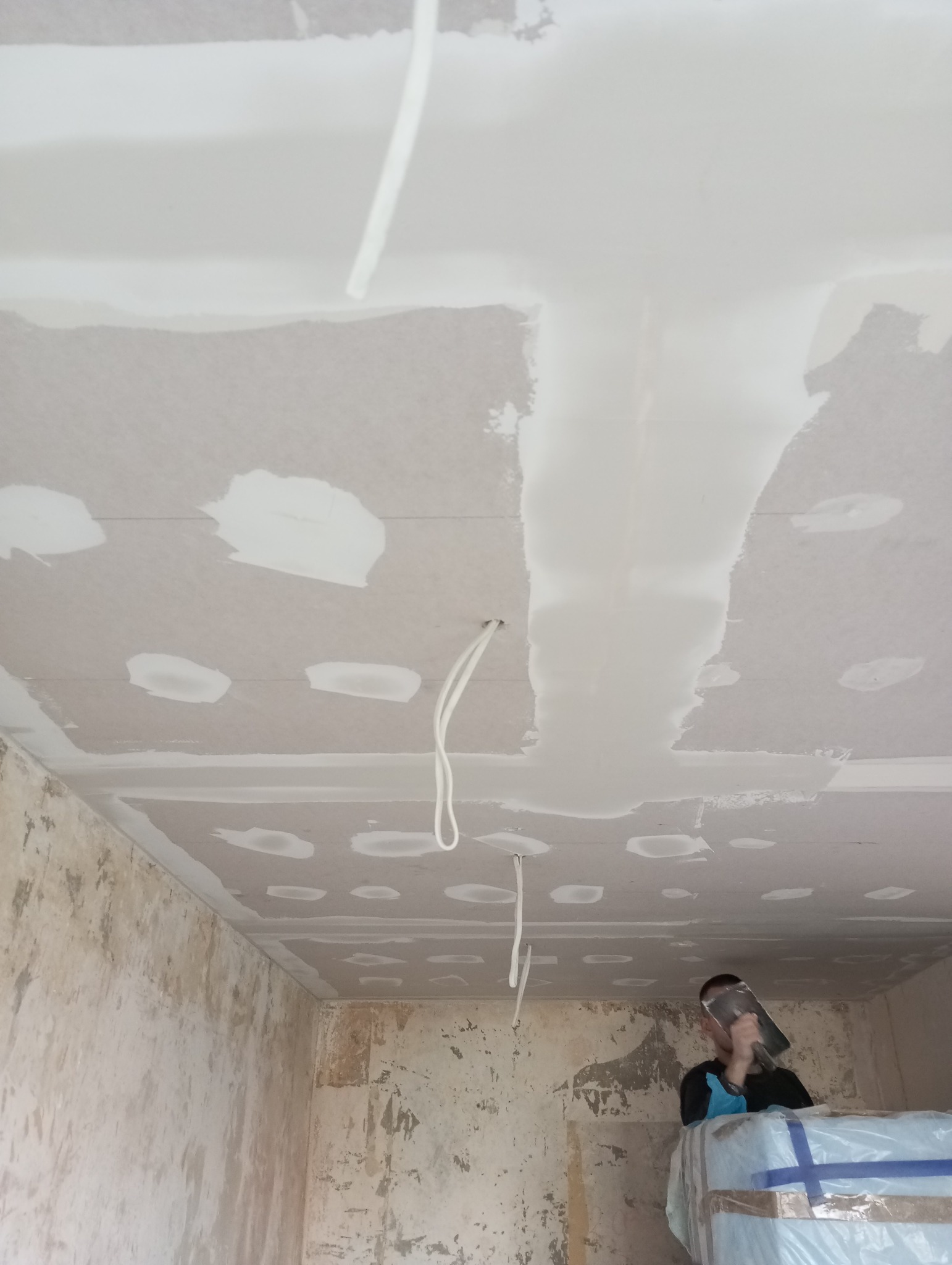 Pracownik szpachluje sufit z płyt kartonowo-gipsowych, widoczne kable wystające z sufitu i ściany z resztkami tapet.