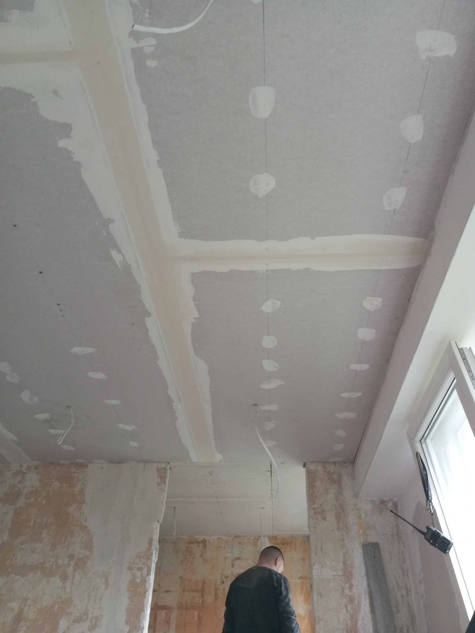 Sufit z płyt kartonowo-gipsowych w trakcie montażu, widoczne szpachlowane łączenia i otwory po wkrętach, ściany w stanie surowym, fragment okna i sylwetka pracownika.