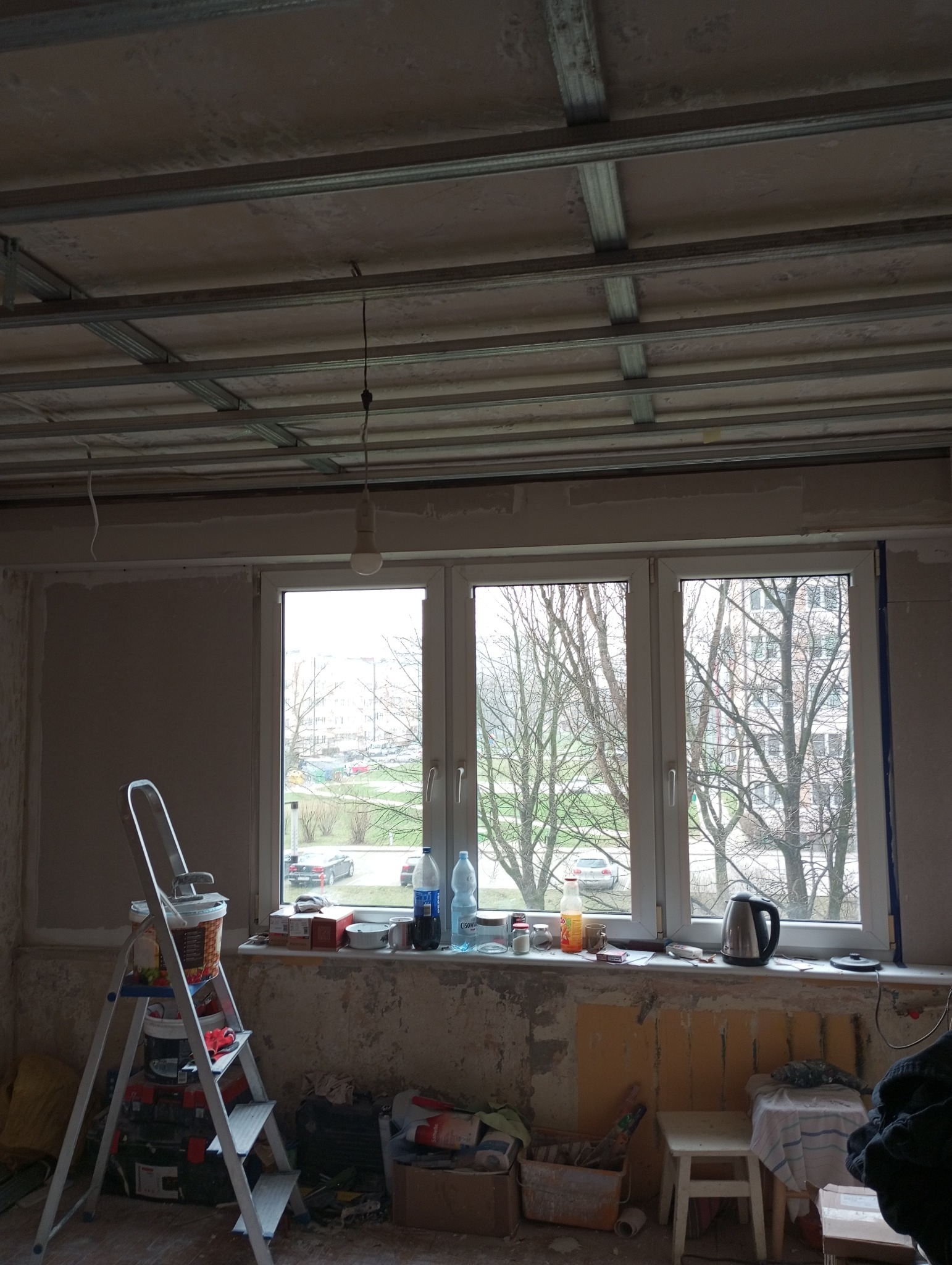 Wnętrze w trakcie remontu: widoczna drabina z farbami, okno z widokiem na drzewa, sufit z metalową konstrukcją pod płyty gipsowe, ściany częściowo wyszpachlowane, bałagan na parapecie i podłodze.