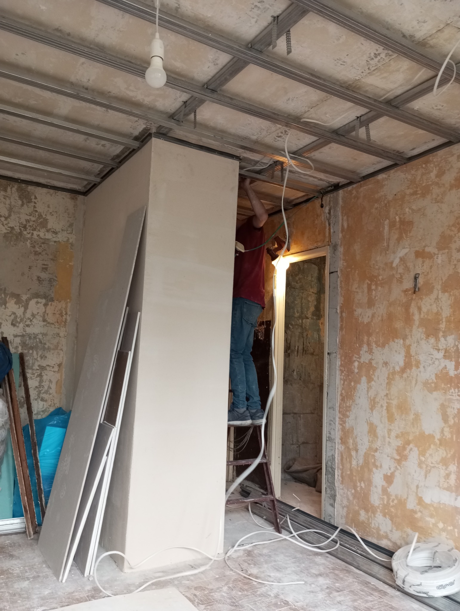 Montaż płyty karton gips na metalowym stelażu sufitowym w pomieszczeniu z częściowo otynkowanymi ścianami, widoczna drabina i zwisające kable elektryczne.