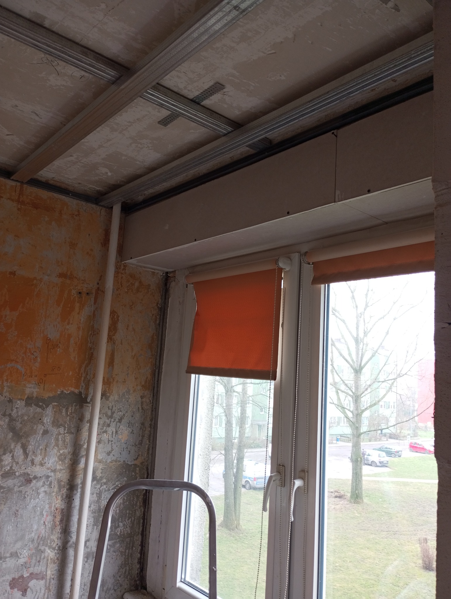 Montaż metalowej konstrukcji sufitu podwieszanego z płyt karton gips, widoczne profile i mocowania, ściany w stanie surowym z resztkami farby, okno z pomarańczową roletą.