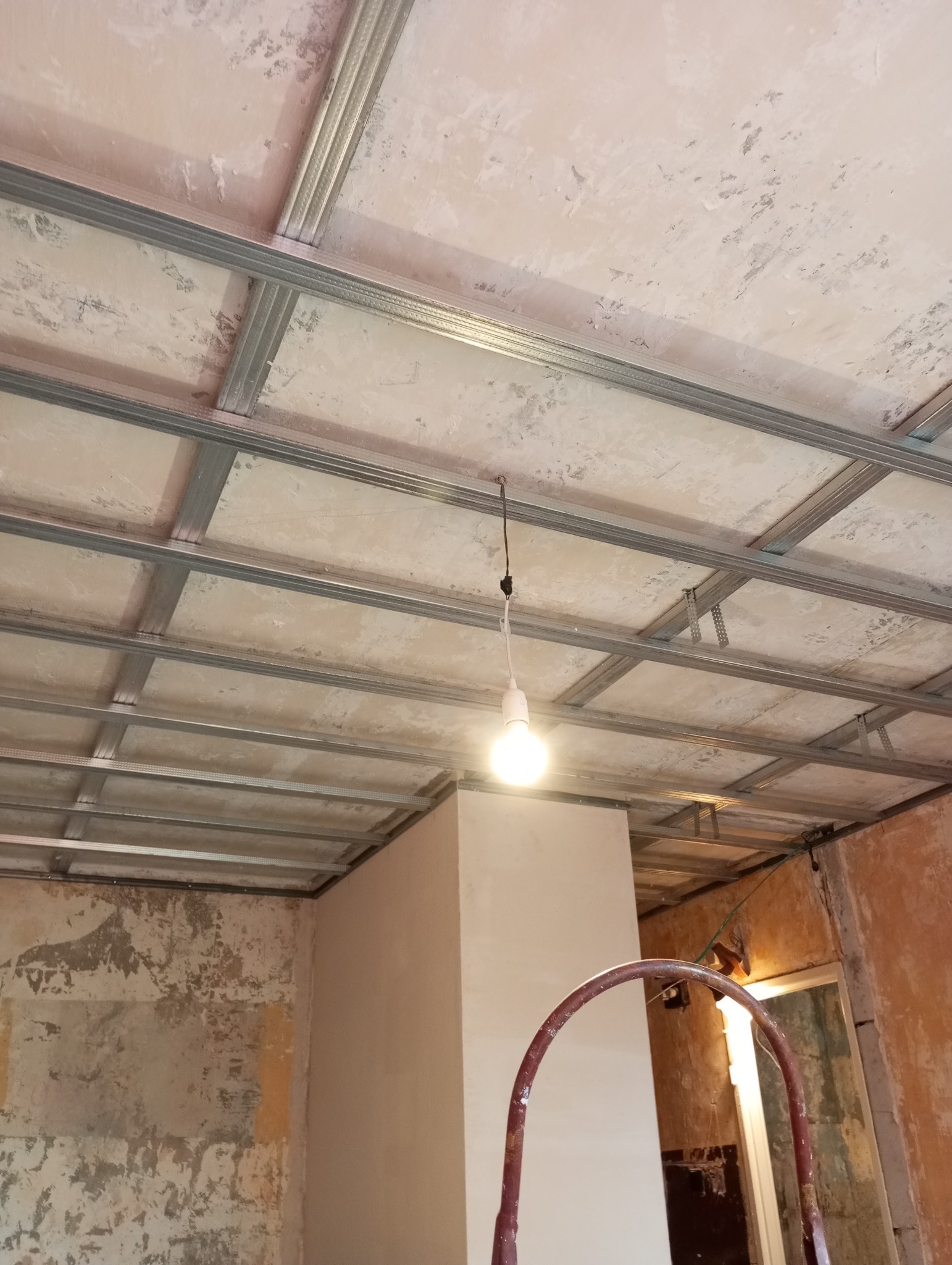 Niedokończony sufit podwieszany z metalowym stelażem i prowizorycznym oświetleniem żarówką, częściowo otynkowane ściany w tle, widoczny fragment drabiny malarskiej.