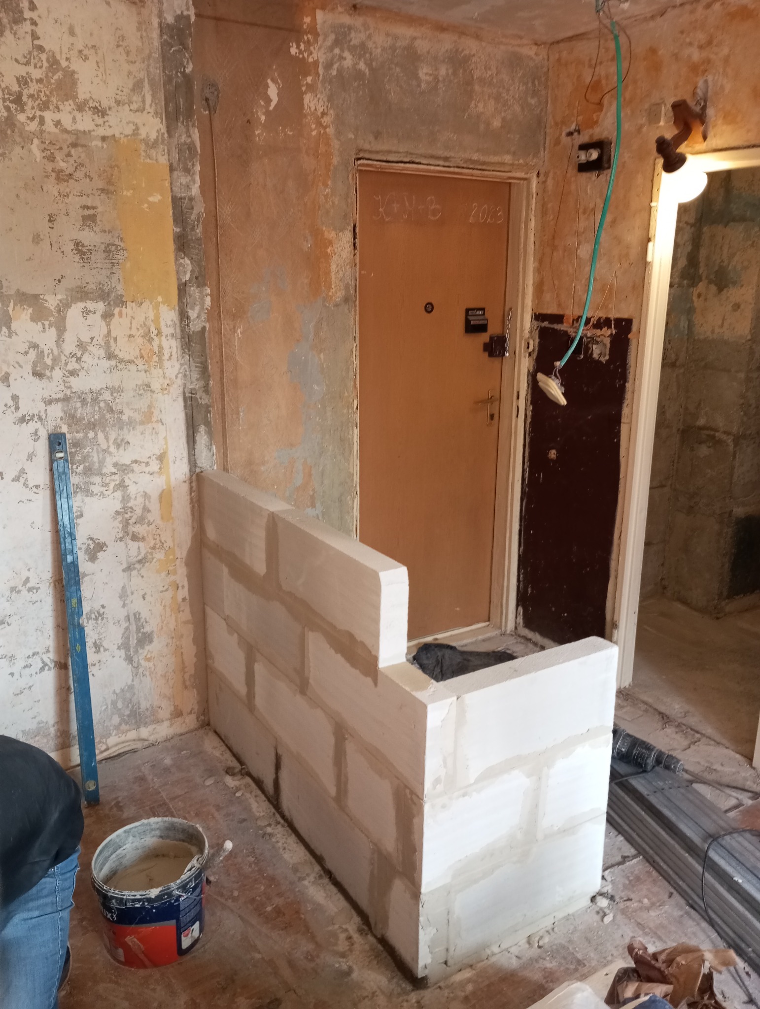 Wnętrze w trakcie remontu: fragment ściany z surowymi blokami, poziomica oparta o ścianę, wiadro z zaprawą, widoczne ślady starej farby i instalacji elektrycznej.