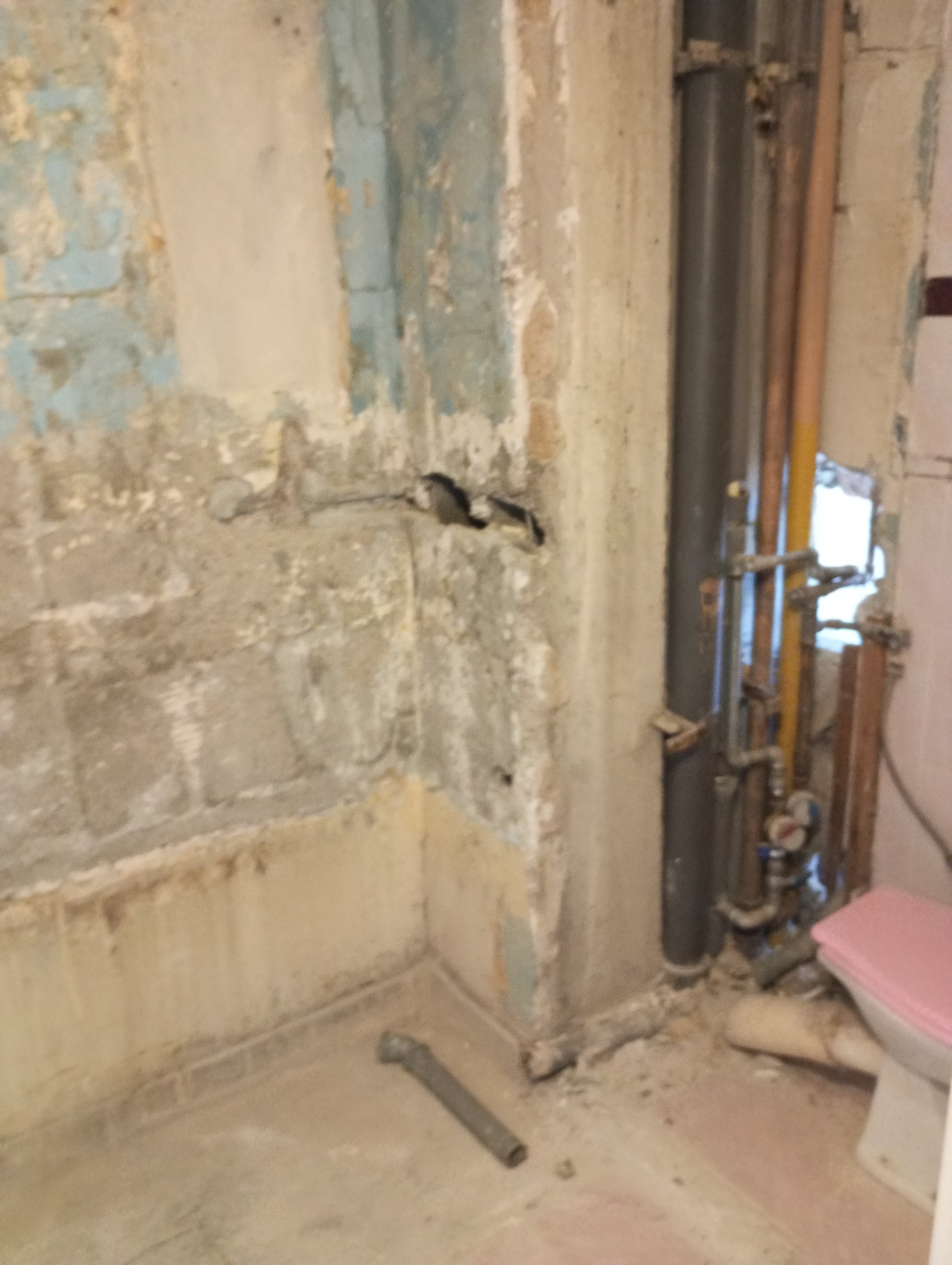 Zdjęcie przedstawia wnętrze łazienki w trakcie remontu, z widocznymi fragmentami starej, zniszczonej ściany z pozostałościami farby, odsłoniętymi rurami i instalacją wodną oraz fragmentem toalety...