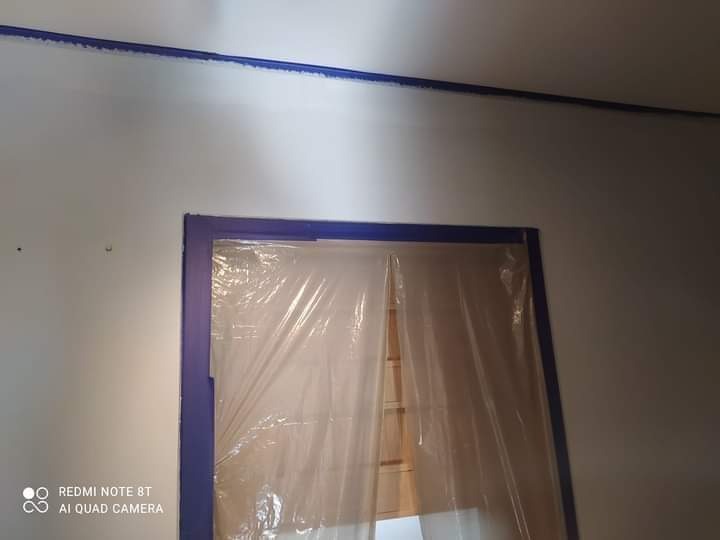 Przygotowanie do malowania: ściana zabezpieczona taśmą malarską przy suficie i wokół drzwi, drzwi zakryte folią ochronną.