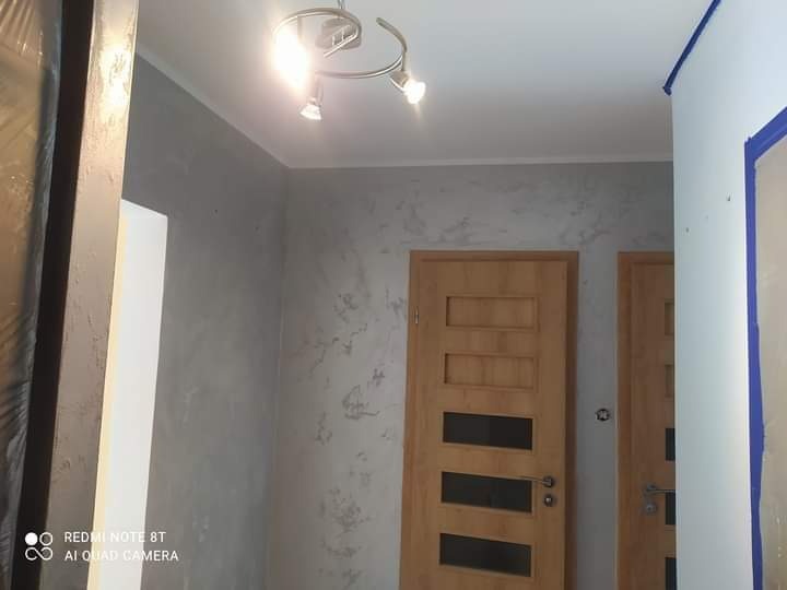 Ściana w pokoju wykończona dekoracyjną szarą farbą strukturalną, widoczne dwie drewniane drzwi z przeszkleniami, oświetlenie sufitowe z trzema reflektorami.