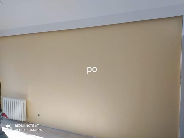 Ściana pomalowana na beżowo z białym grzejnikiem i listwą przysufitową, widoczny napis 'po' na ścianie.