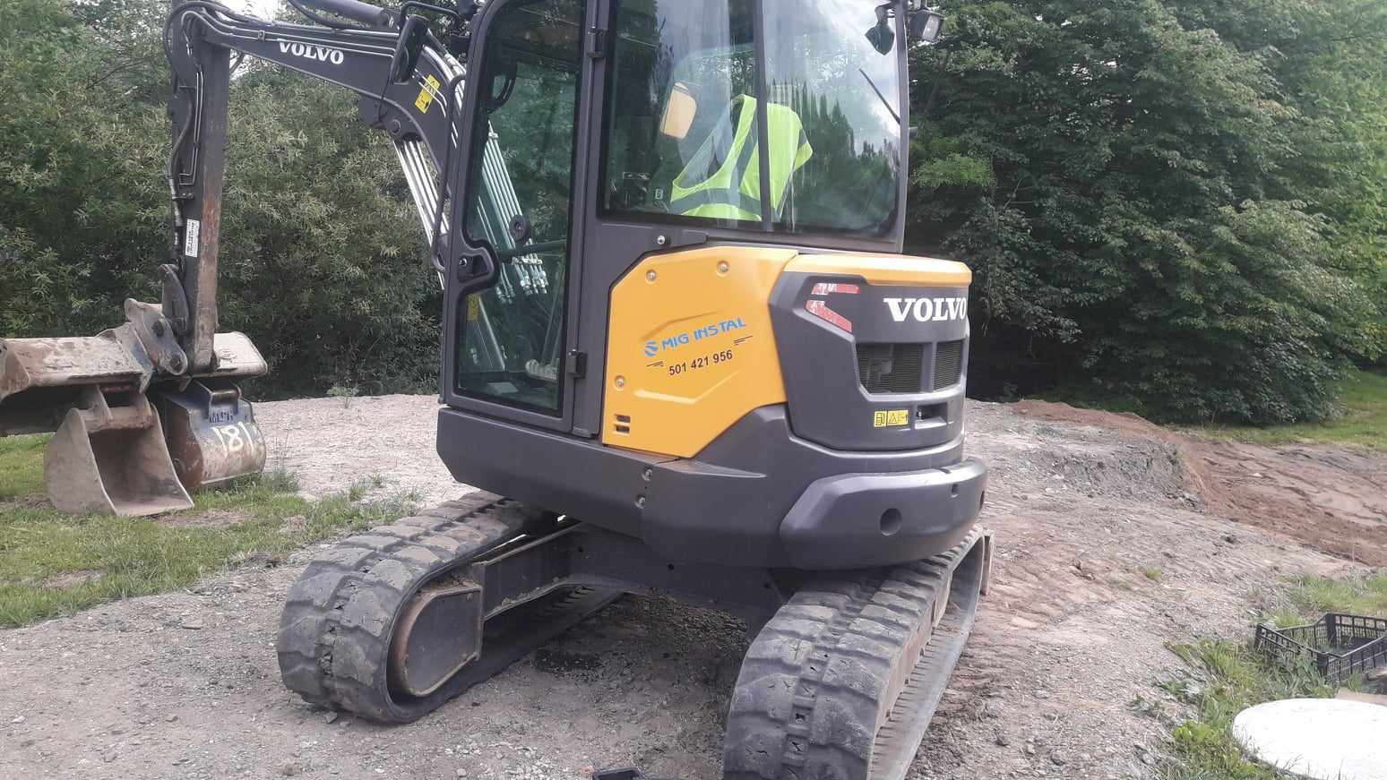 Żółto-szara koparka Volvo z łyżką, widoczna na terenie budowy z wykopaną ziemią w tle, z numerem telefonu firmy na obudowie.