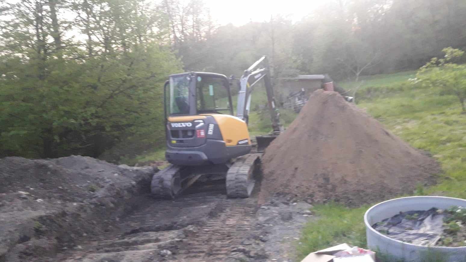 Żółto-szara koparka Volvo EC160 na gąsienicach, stojąca obok wykopu i pryzmy ziemi na zewnątrz, w tle drzewa.