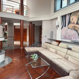 Katarzyna Rychlik Nieruchomości - Przestronny salon z kominkiem obudowanym marmurem, parkietem z ciemnego drewna, beżową sofą modułową i nowoczesnym obrazem na ścianie.