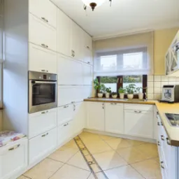 Jasna kuchnia w stylu klasycznym z białymi szafkami, drewnianym blatem, roślinami w doniczkach na parapecie i beżowymi płytkami podłogowymi z dekoracyjnym pasem.