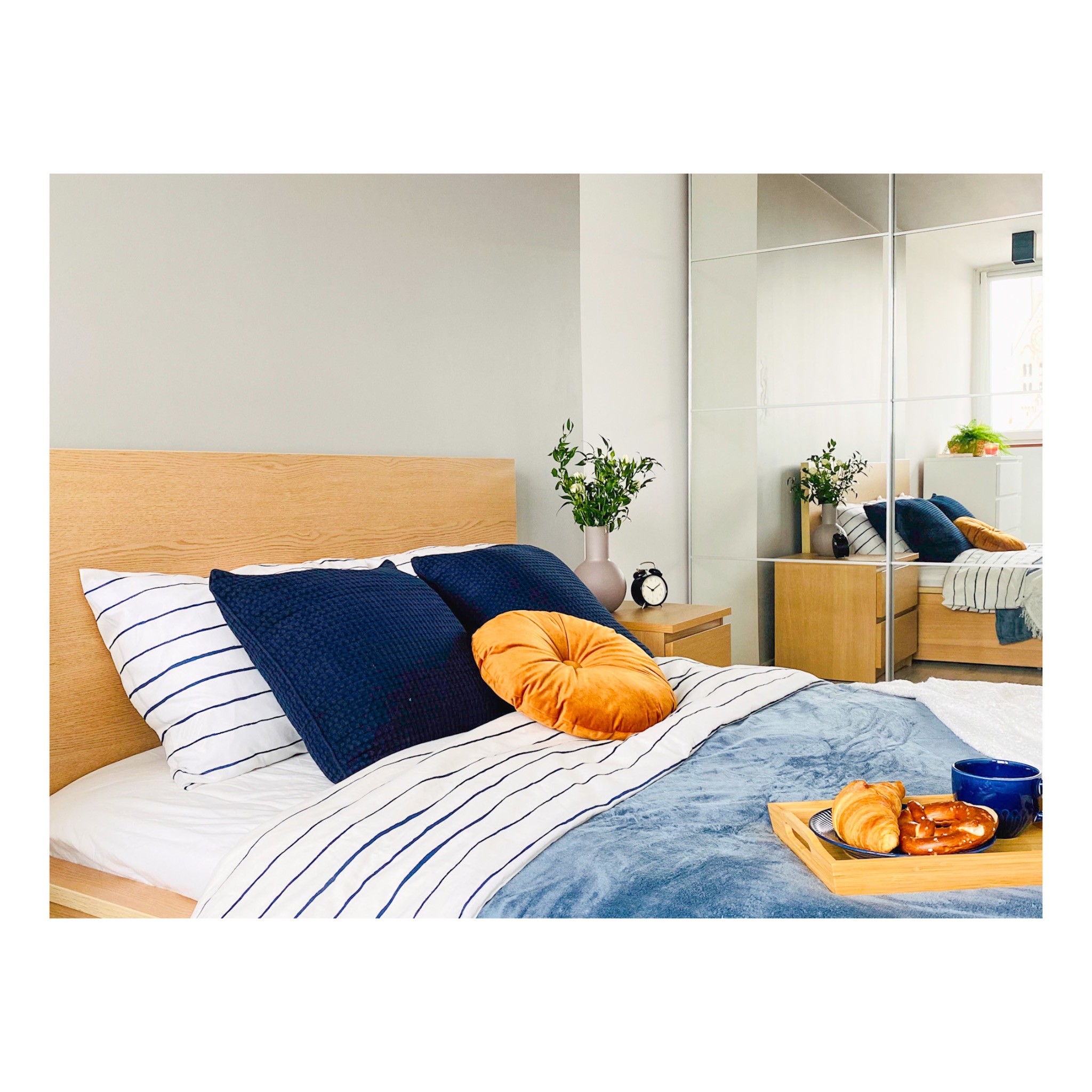 Jasne drewniane łóżko z niebieskimi poduszkami, okrągłą pomarańczową poduszką, pościelą w paski i śniadaniem na tacy, odbijające się w lustrzanych drzwiach szafy.