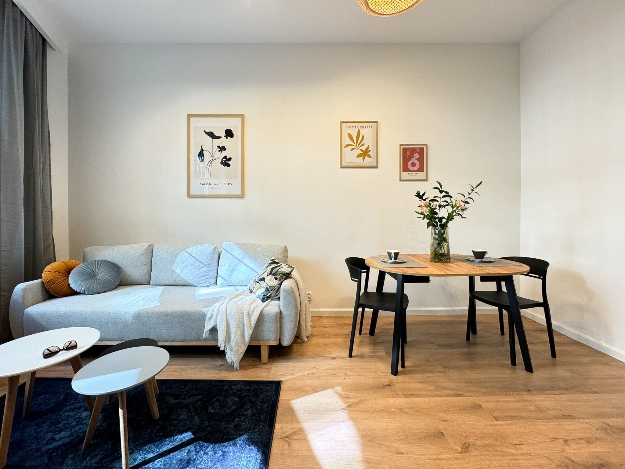 Jasne wnętrze mieszkania z drewnianą podłogą, szarą sofą z dekoracyjnymi poduszkami, okrągłym stołem z czarnymi krzesłami i trzema obrazkami na ścianie.