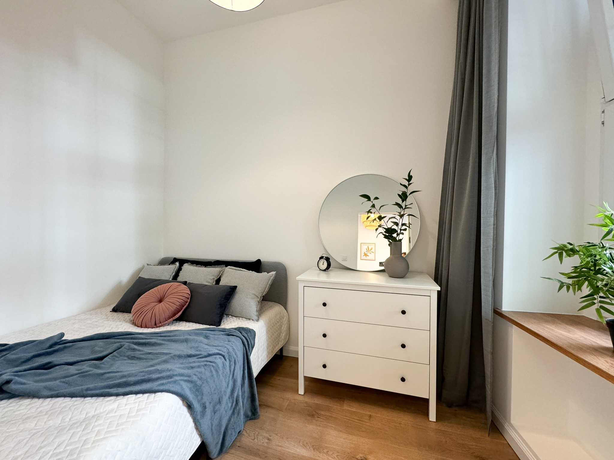 Jasna sypialnia z łóżkiem, komodą z okrągłym lustrem, szarą zasłoną i drewnianą podłogą, w lustrze odbija się plakat z motywem roślinnym.