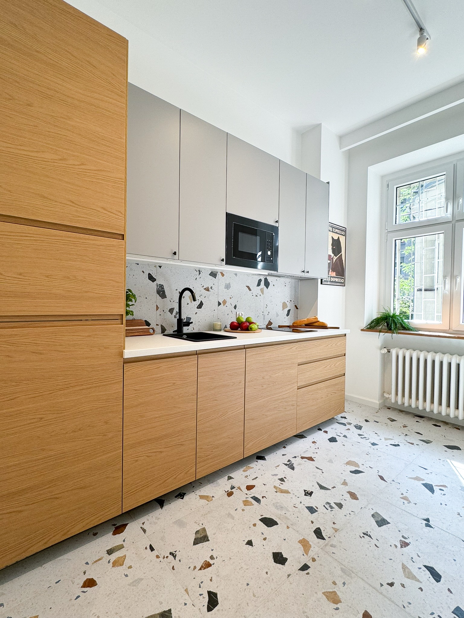 Nowoczesna kuchnia z drewnianymi i szarymi szafkami, blatem z czarnym zlewem i baterią, oraz podłogą w stylu lastryko. Na ścianie plakat z czarnym kotem w garniturze.