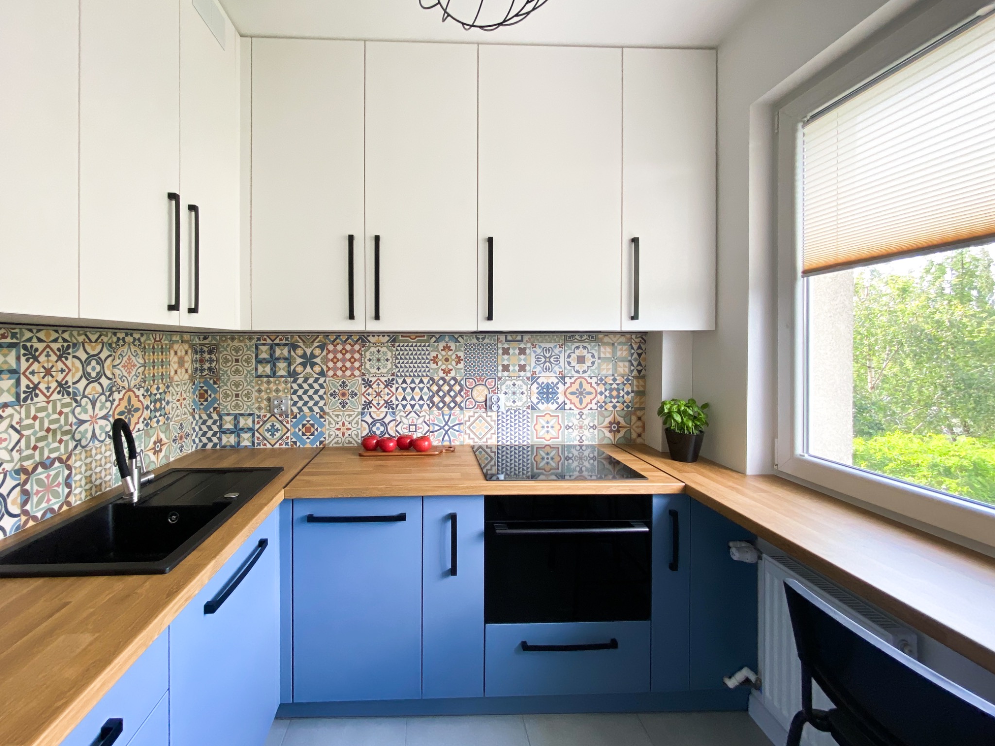 Nowoczesna kuchnia w Katowicach z błękitnymi szafkami dolnymi, białymi górnymi, drewnianym blatem i kolorowymi płytkami backsplash, z czarnym zlewem i trzema czerwonymi jabłkami na desce.