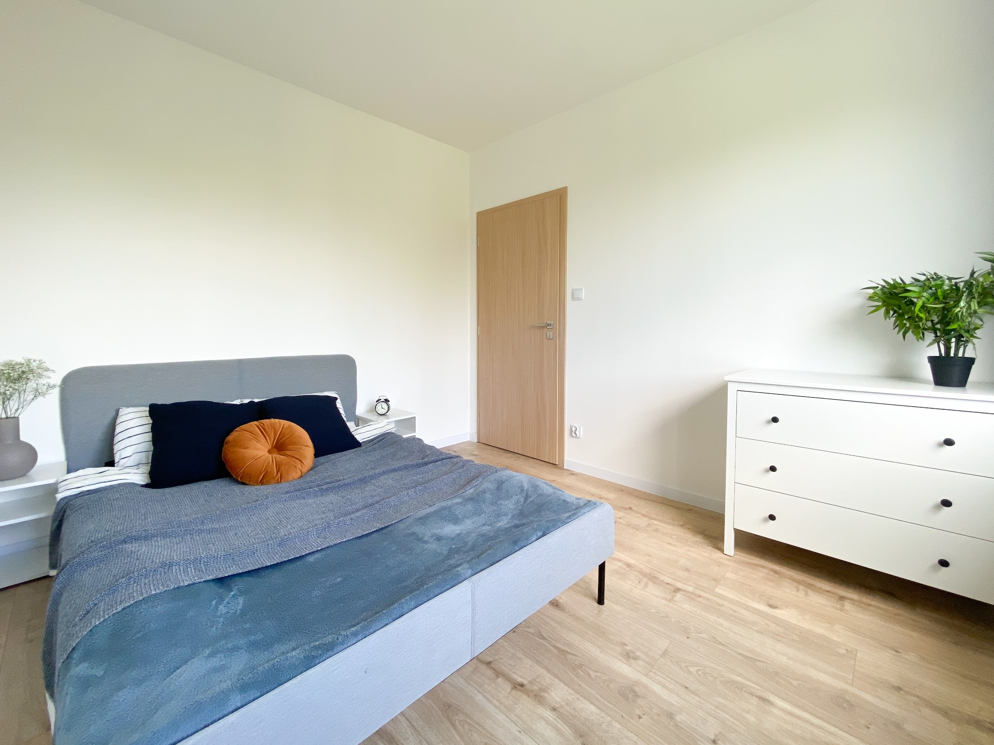 Jasne wnętrze sypialni z łóżkiem z szarą ramą, niebieską narzutą i okrągłą pomarańczową poduszką, białą komodą z rośliną doniczkową i drewnianą podłogą.