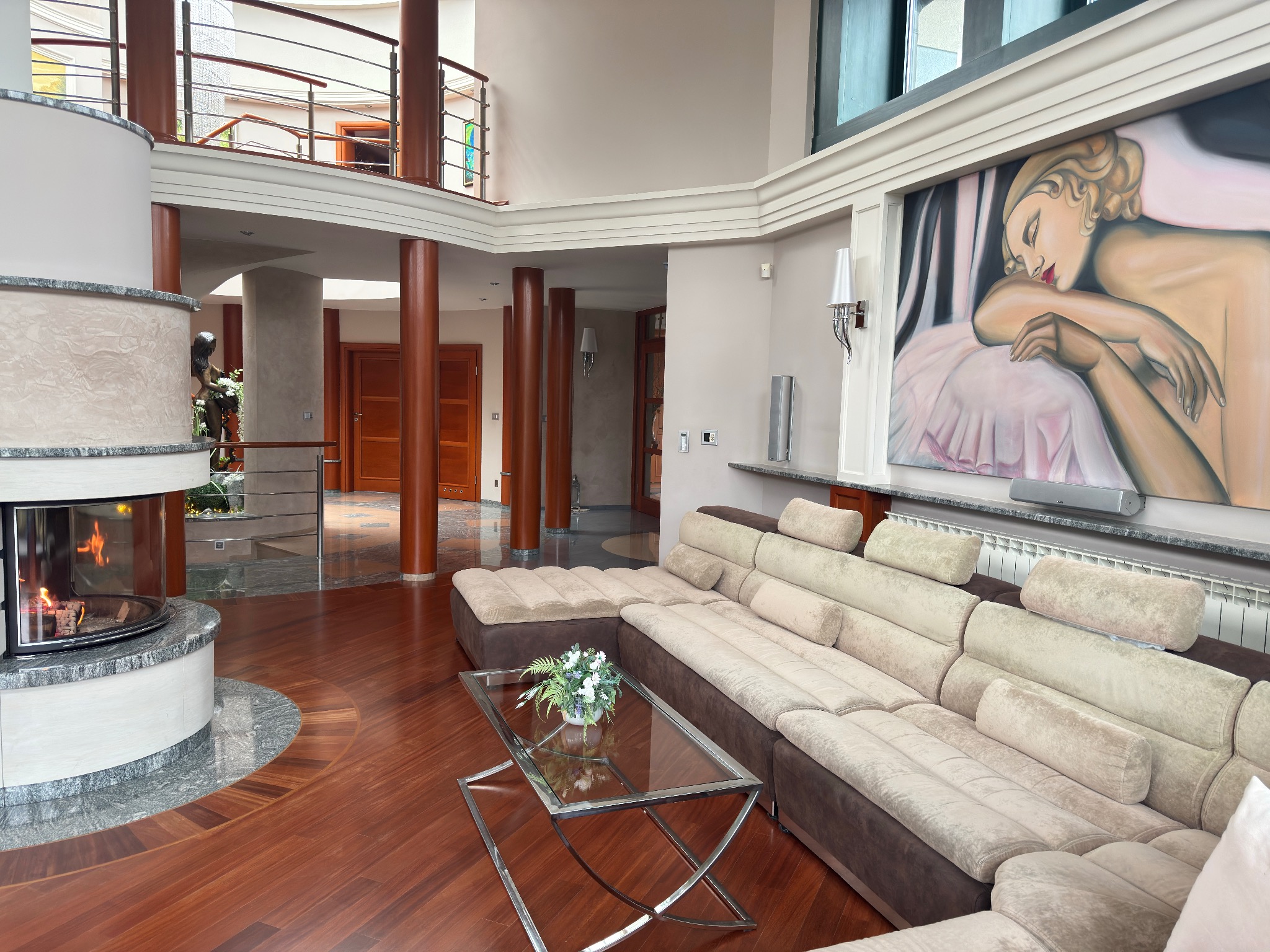 Przestronny salon z kominkiem obudowanym marmurem, parkietem z ciemnego drewna, beżową sofą modułową i nowoczesnym obrazem na ścianie.