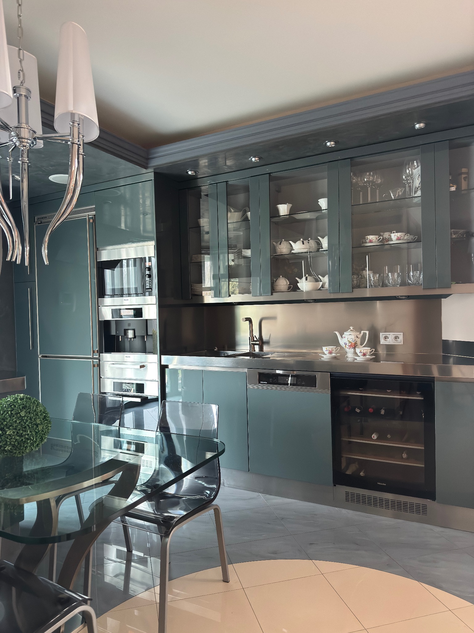 Nowoczesna kuchnia w odcieniach szarości i błękitu z zabudową meblową, szklanymi witrynami, sprzętem AGD marki Miele i szklanym stołem z krzesłami.