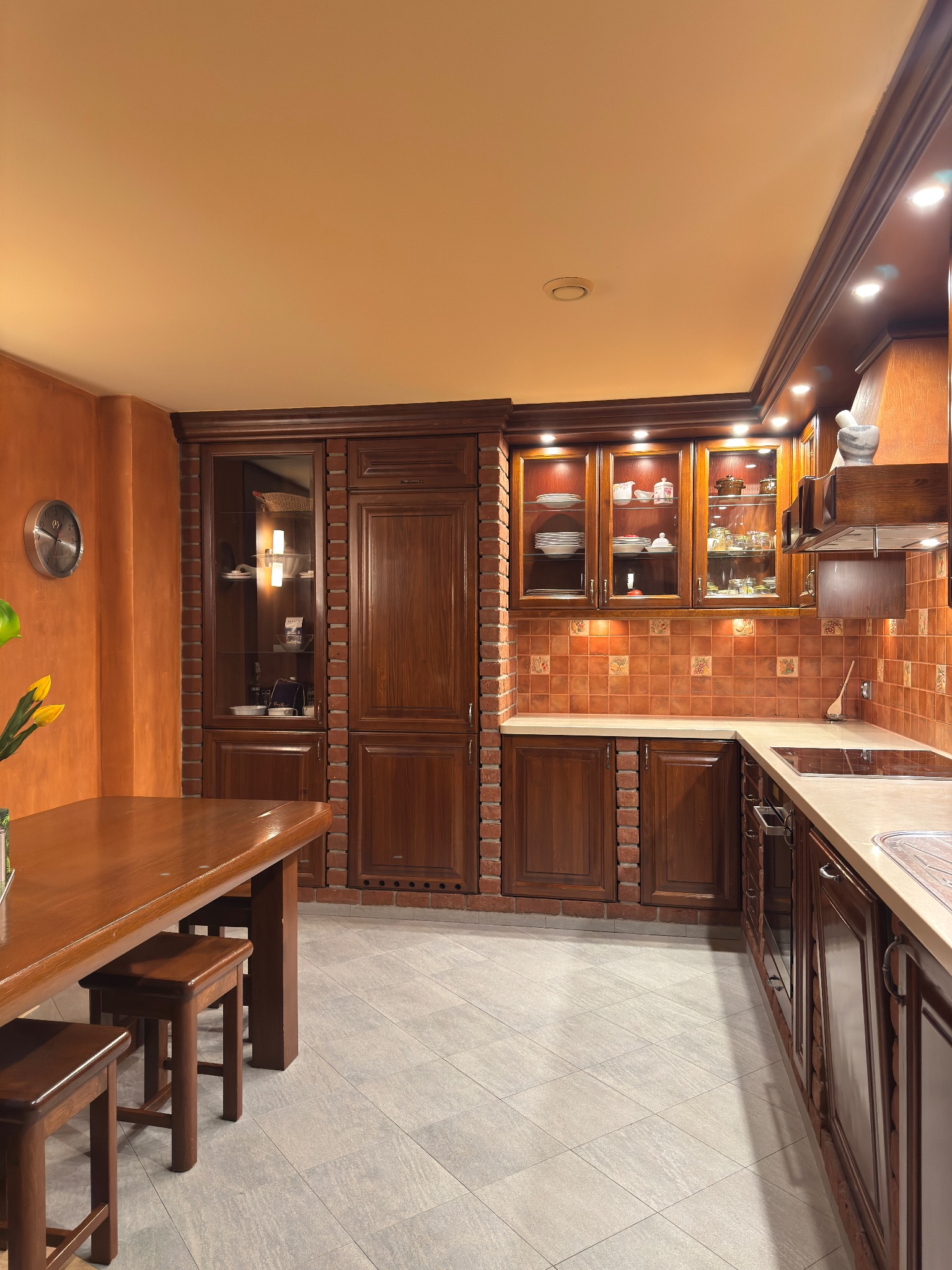 Rustykalna kuchnia z drewnianymi szafkami, ceglanymi akcentami, oświetleniem punktowym i stołem z krzesłami w tle.