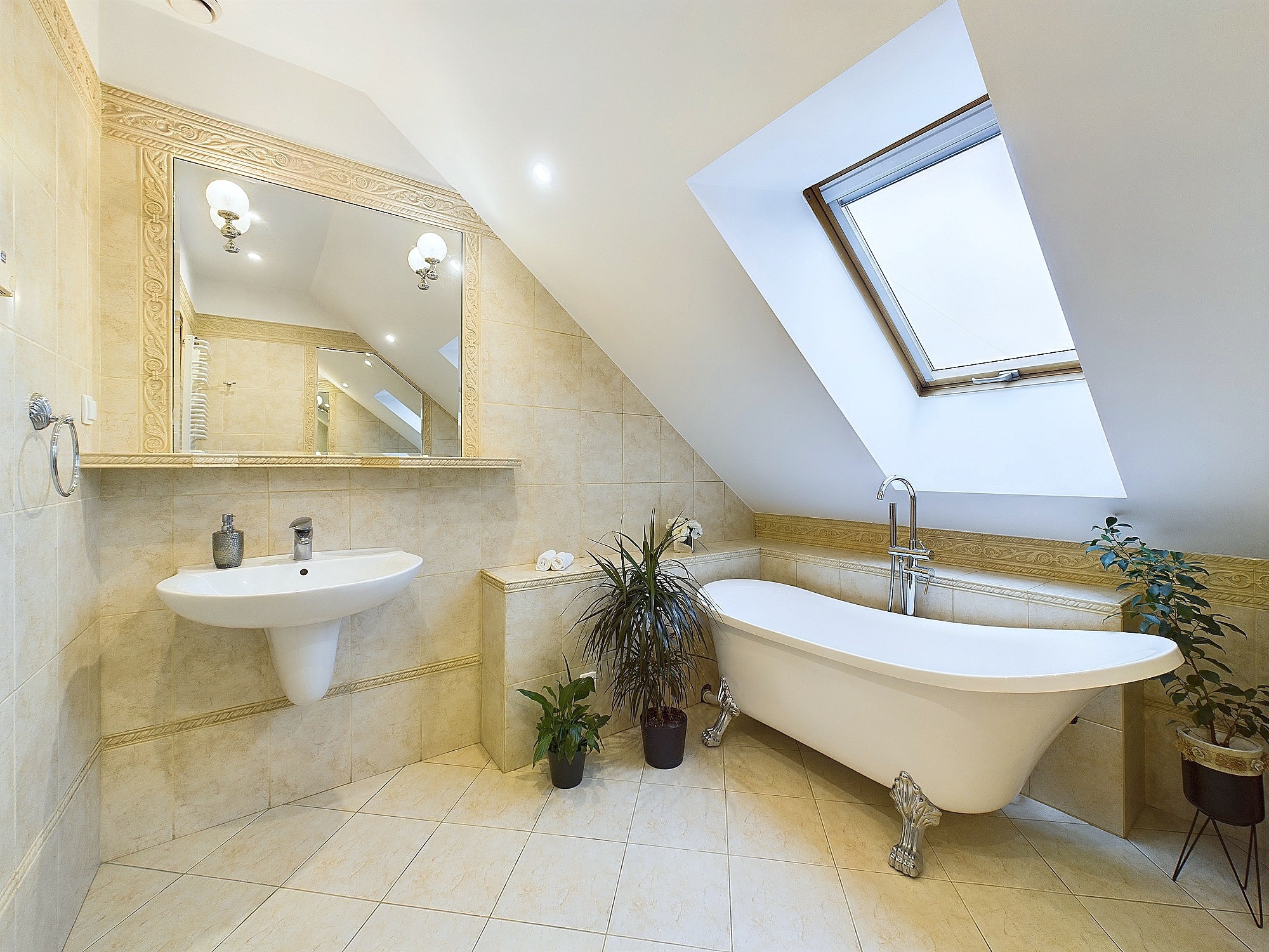 Jasna łazienka na poddaszu z wanną na lwich nóżkach, umywalką na postumencie i oknem dachowym, wykończona beżowymi płytkami z dekoracyjnym ornamentem.