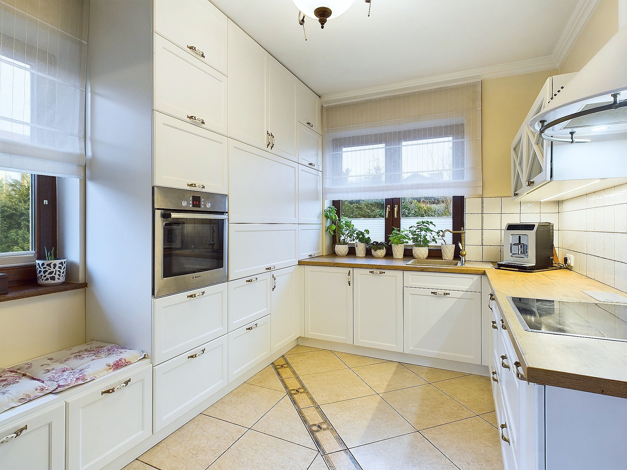 Jasna kuchnia w stylu klasycznym z białymi szafkami, drewnianym blatem, roślinami w doniczkach na parapecie i beżowymi płytkami podłogowymi z dekoracyjnym pasem.