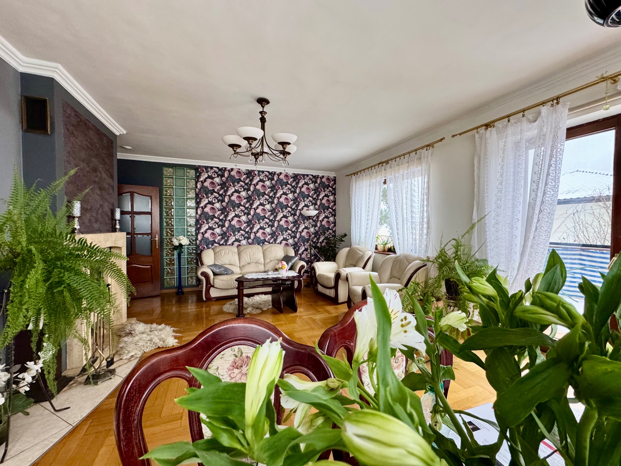 Jasny salon z parkietem, kremowymi sofami i fotelami, kwiatową tapetą, żyrandolem i roślinami doniczkowymi, widziany zza bukietu białych lilii na pierwszym planie.