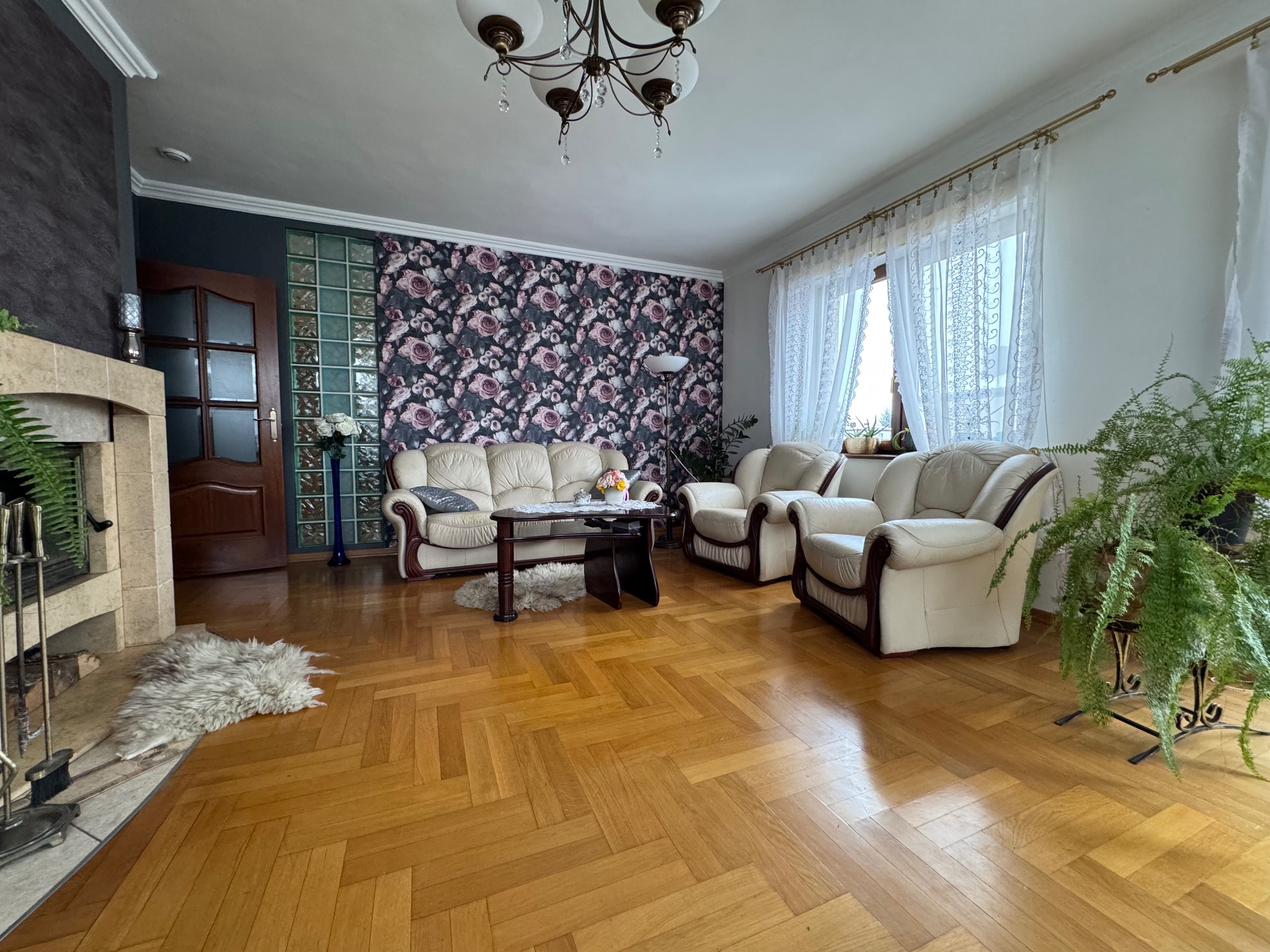 Jasny salon z parkietem w jodełkę, kremowymi skórzanymi sofami, kominkiem z elementami kamienia i ścianą w kwiatowy wzór, oświetlony przez żyrandol z kryształkami.