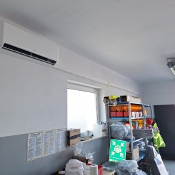 KlimaGold TOMASZ GUCWA - Biała jednostka klimatyzacyjna Mistral zamontowana na ścianie w pomieszczeniu warsztatowym, widoczne instrukcje BHP, półki z akcesoriami i otwarte drzwi garażowe.