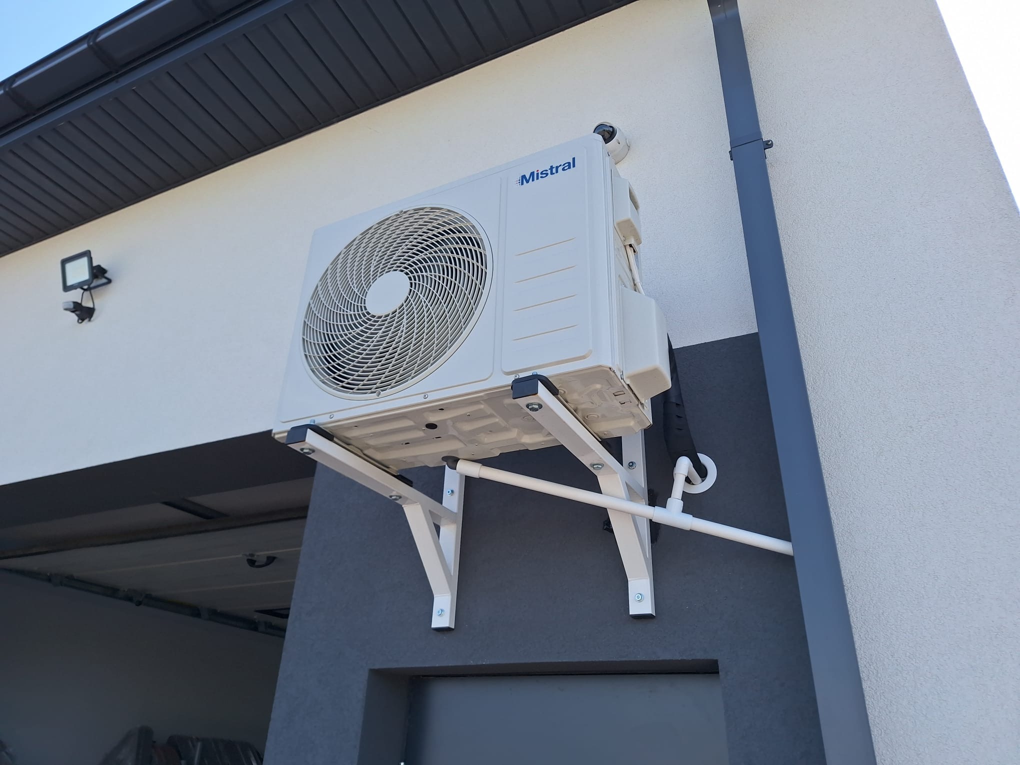 Zewnętrzna jednostka klimatyzacji Mistral zamontowana na białym wsporniku na elewacji budynku z szarą dolną częścią i białą górną częścią, widoczna rynna i oświetlenie zewnętrzne.