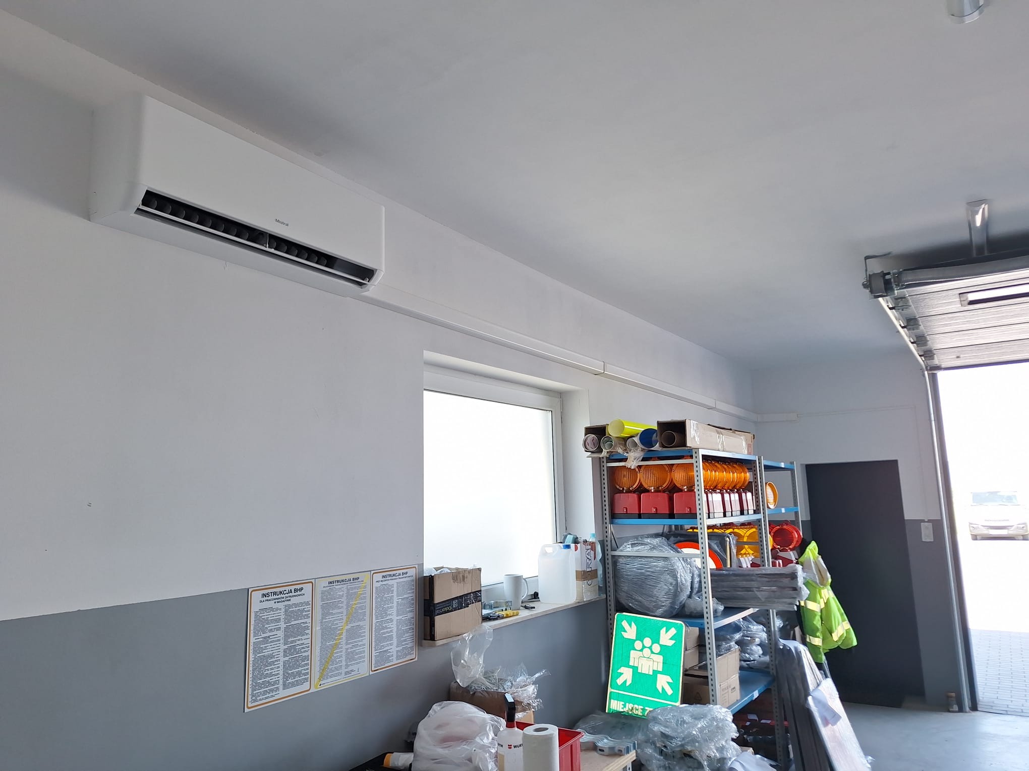 Biała jednostka klimatyzacyjna Mistral zamontowana na ścianie w pomieszczeniu warsztatowym, widoczne instrukcje BHP, półki z akcesoriami i otwarte drzwi garażowe.