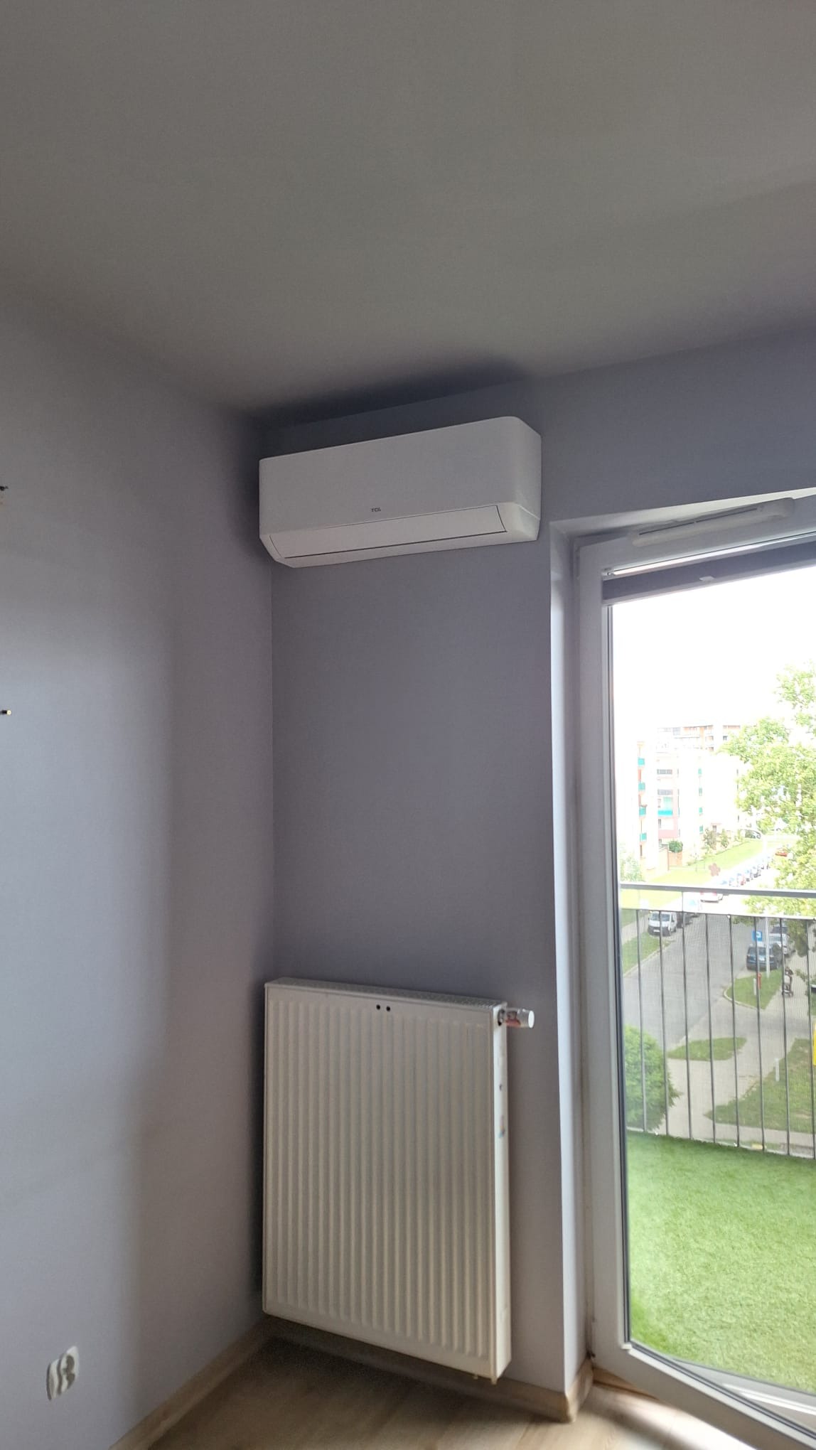 Biały klimatyzator TCL zamontowany pod sufitem w szarym pokoju, obok okna balkonowego i grzejnika.