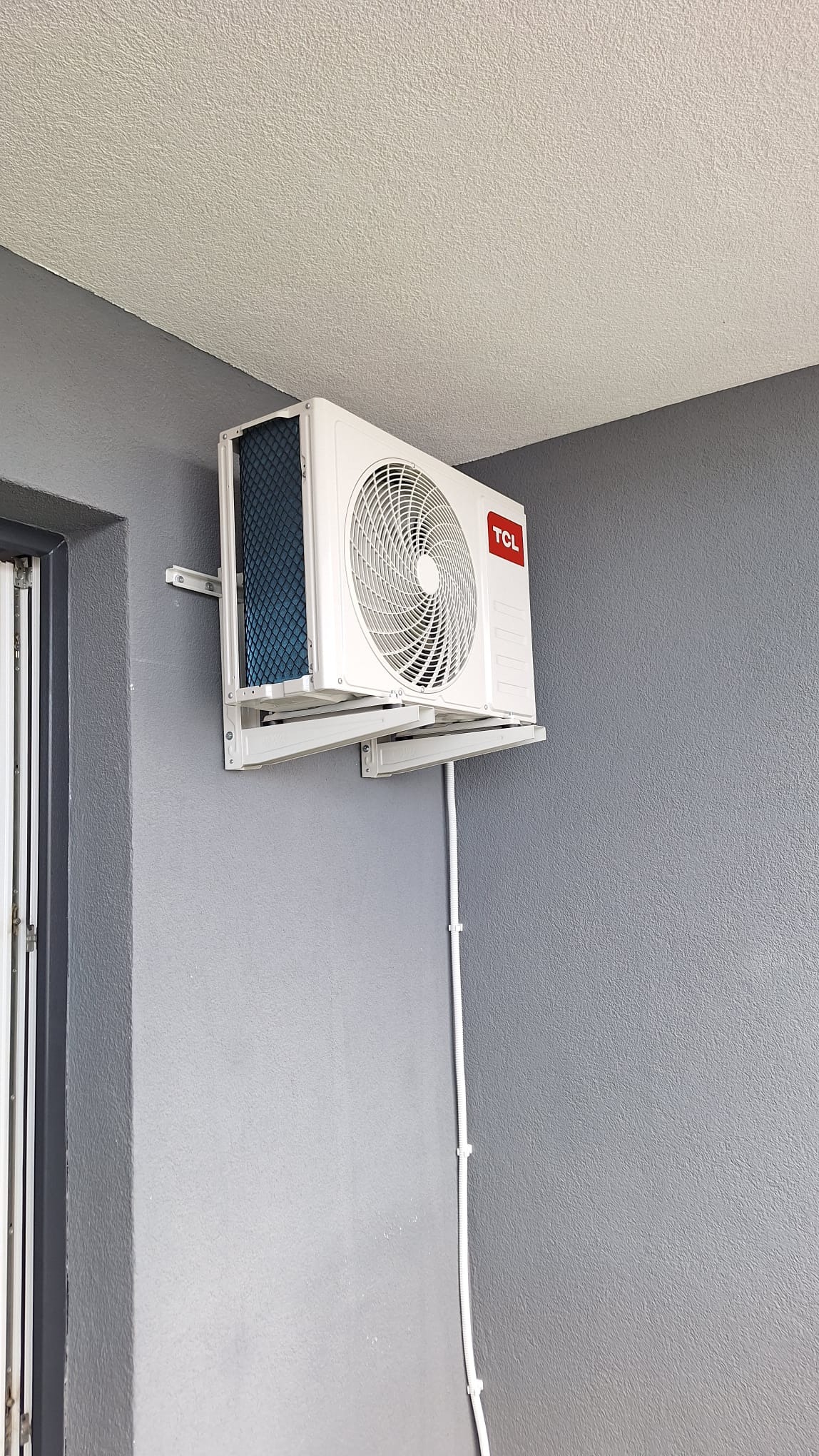 Biała jednostka klimatyzacyjna TCL zamontowana na szarej ścianie z widocznym kablem w plastikowej osłonie.