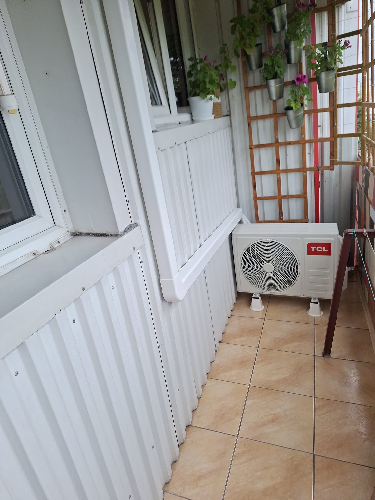 Biała jednostka zewnętrzna klimatyzatora TCL zamontowana na balkonie z brązowymi płytkami podłogowymi, na tle białej ściany z panelami i dekoracyjnej drabinki z wiszącymi doniczkami z kwiatami.