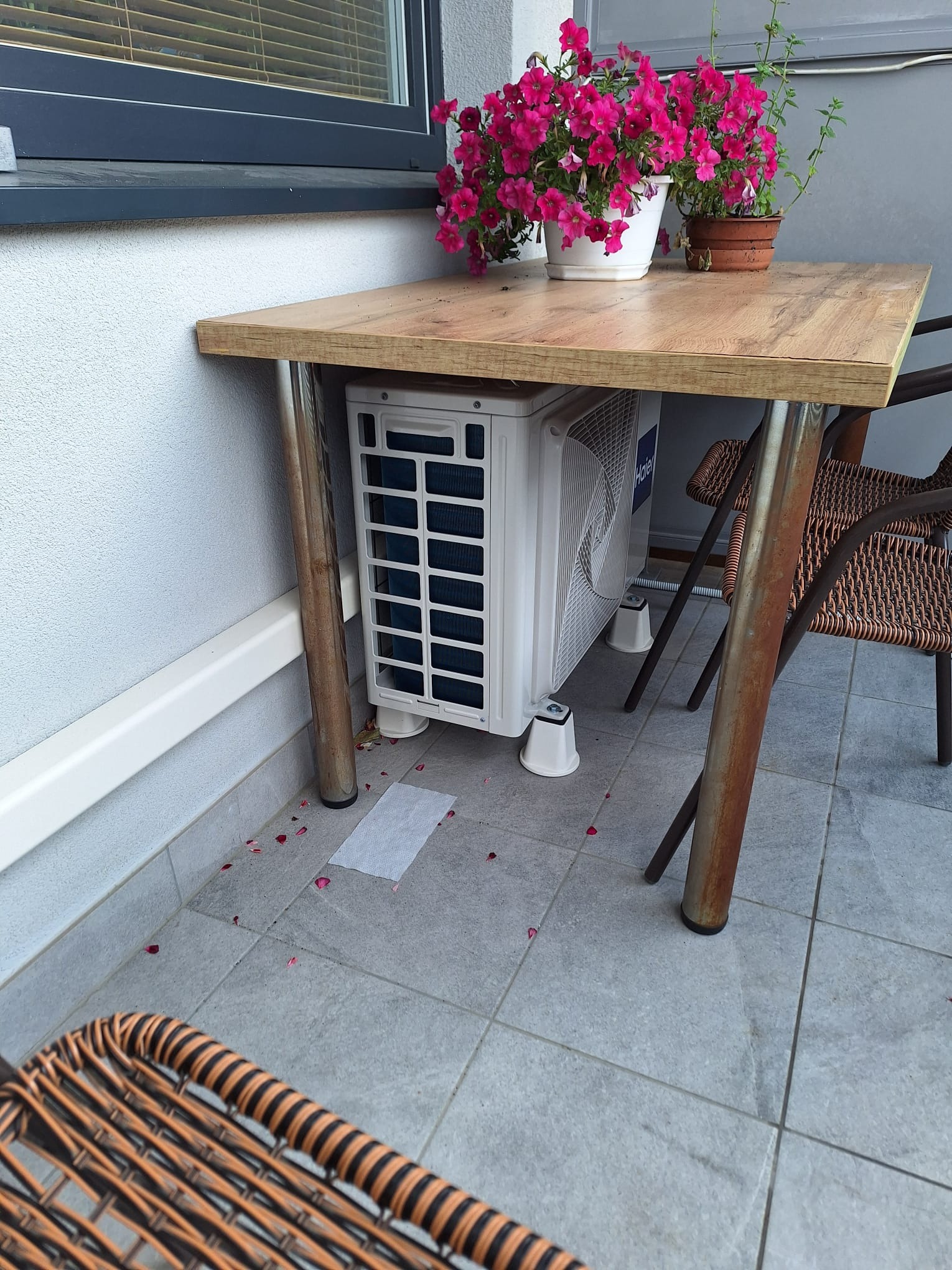 Kompaktowy klimatyzator zewnętrzny marki Haier, umieszczony pod drewnianym stołem na balkonie, z doniczkami z kwiatami na blacie i wiklinowymi krzesłami obok.