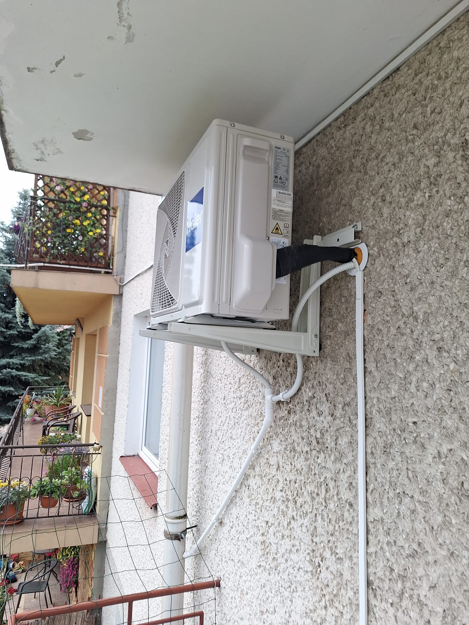 Zewnętrzna jednostka klimatyzatora Haier zamontowana na ścianie budynku z fakturą baranka, widoczne rury odprowadzające skropliny i instalacja elektryczna, balkon z kwiatami w tle.
