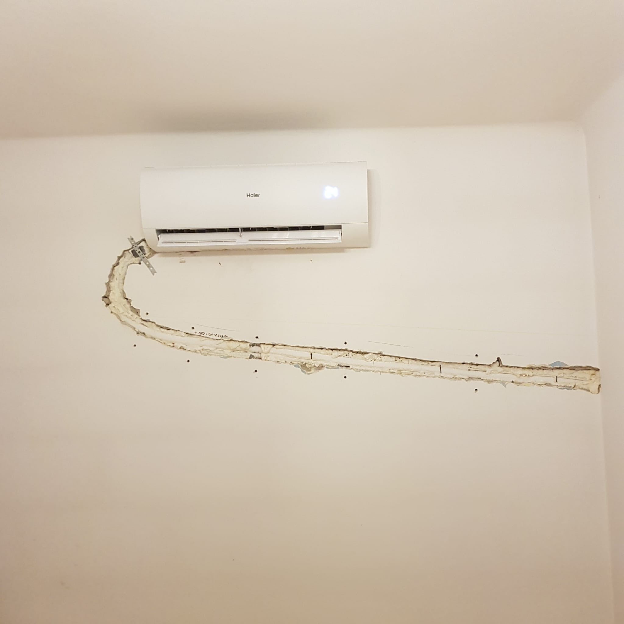 Biały klimatyzator Haier zamontowany na ścianie w trakcie instalacji, widoczne wycięcie w ścianie z przewodami zabezpieczonymi pianką montażową.