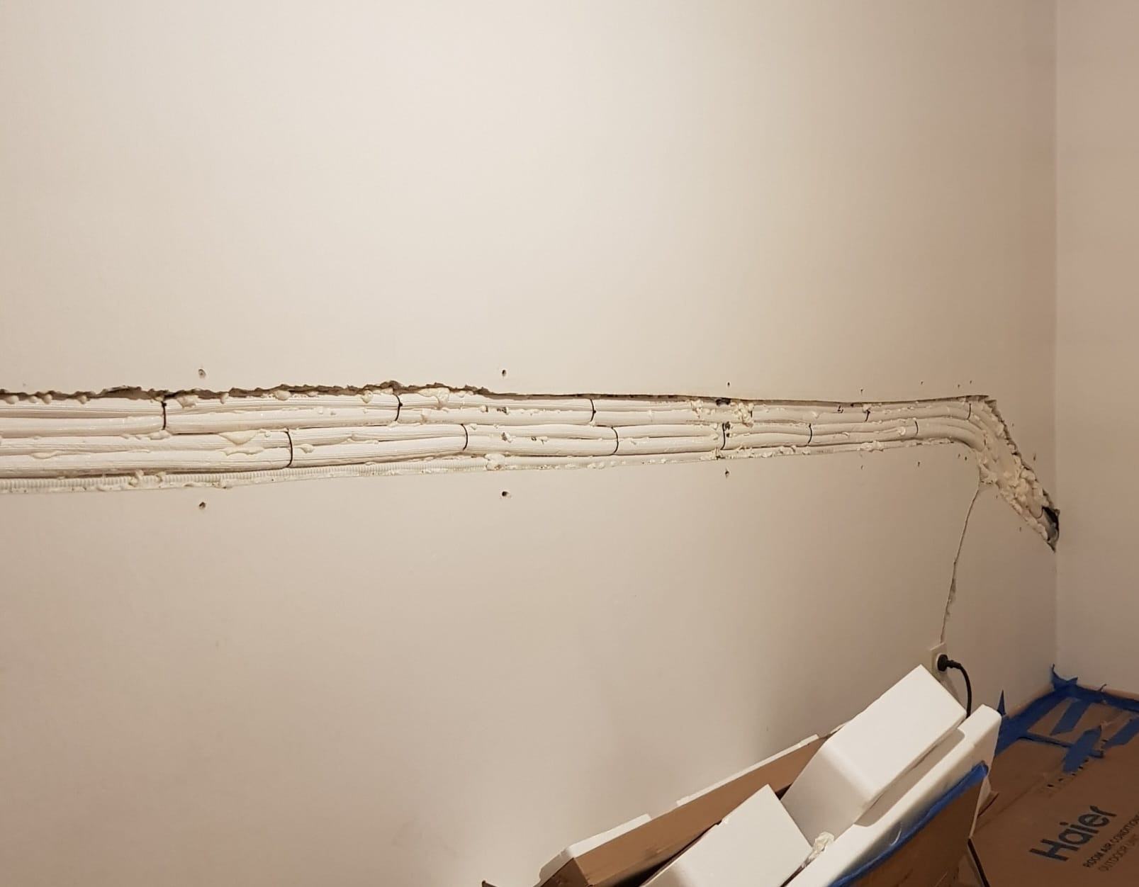 Wnęka w ścianie z ułożonymi rurami instalacyjnymi, zabezpieczonymi pianką montażową i przytwierdzonymi czarnymi opaskami zaciskowymi. Fragment ściany uszkodzony, widoczne pozostałości po pracach...